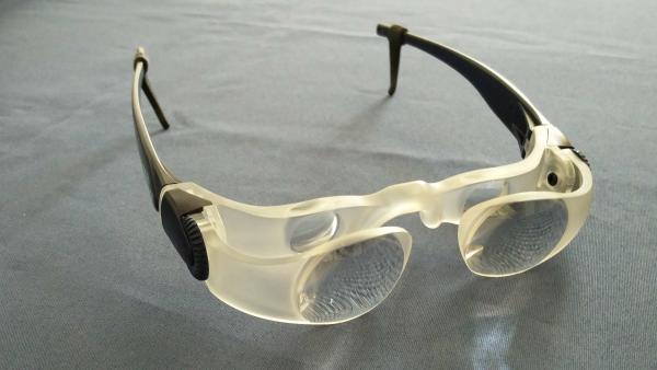 Brille mit Lupengläsern, davor weitere Lupe und Stellrädchen am Brillenbügel